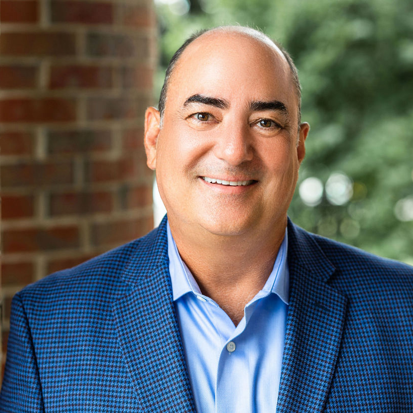 Smiling man in a blue blazer and shirt stands in front of a brick wall and greenery.