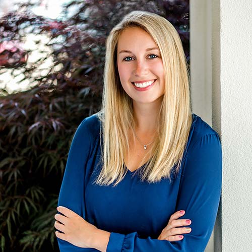 Smiling woman with long blonde hair, wearing a blue top, standing by a wall with arms crossed, outdoor background.