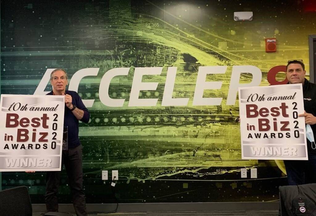 Two people holding 2020 Best in Biz Awards signs in front of a futuristic "ACCELER" background.