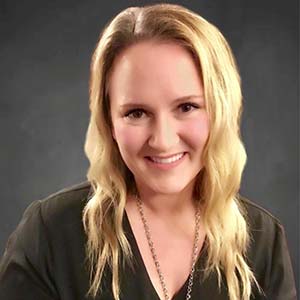 Smiling person with long, wavy blonde hair against a dark background, wearing a dark top and a necklace.