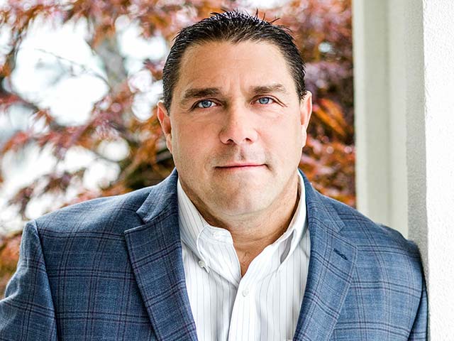 Man in a plaid blazer and white shirt stands outdoors near a wall, with trees in the background.