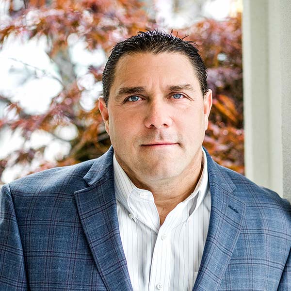 Man in a plaid suit jacket and striped shirt stands in front of autumn foliage.