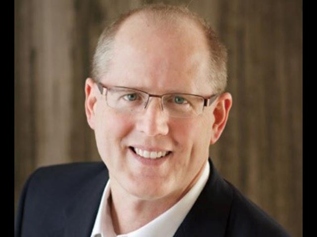 Smiling person with glasses, wearing a suit, posing against a blurred brown background.