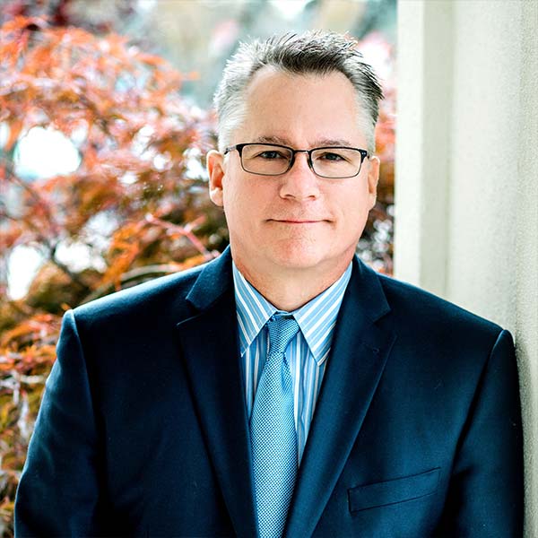 Man in glasses and suit with a blue tie, standing outdoors near a red-leafed tree and a building wall.