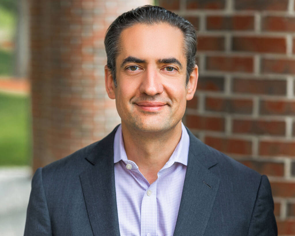 A man in a gray suit and light shirt smiles, standing near a brick wall.