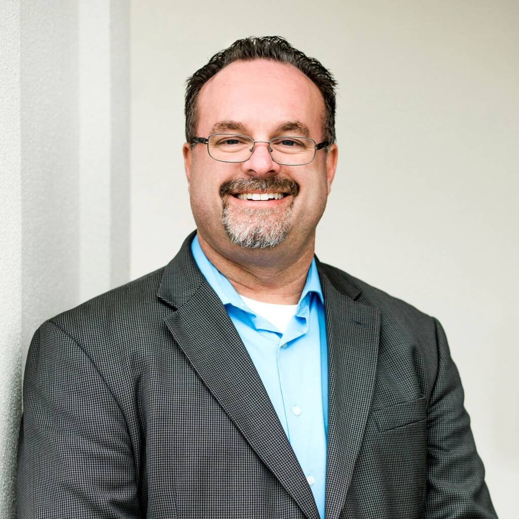Smiling man in glasses wearing a gray suit jacket and blue shirt, standing against a light-colored wall.