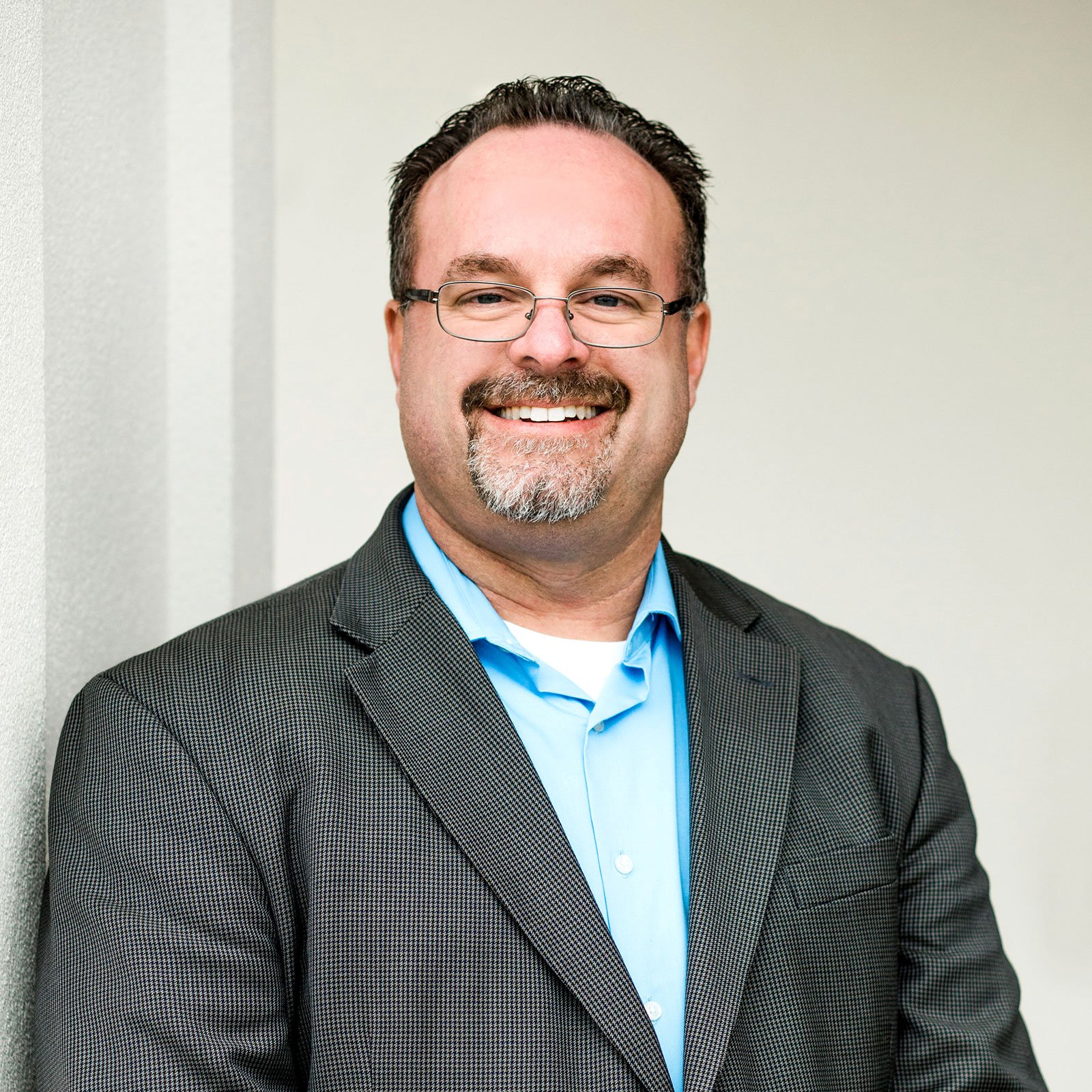 Smiling man in glasses wearing a gray suit jacket and blue shirt, standing against a light-colored wall.