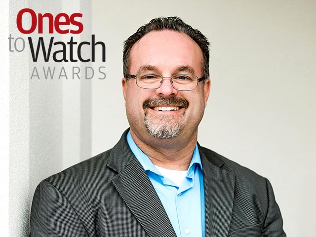 Smiling man in a suit with glasses stands beside "Ones to Watch Awards" text.
