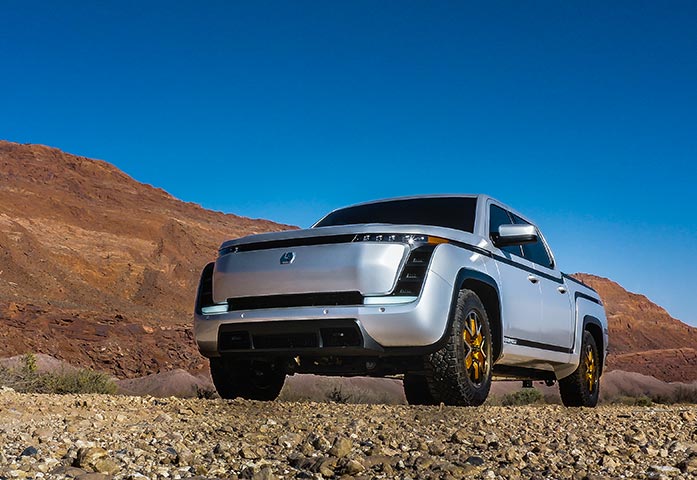 Silver pickup truck with golden rims parked on rocky terrain with red hills and blue sky in the background.