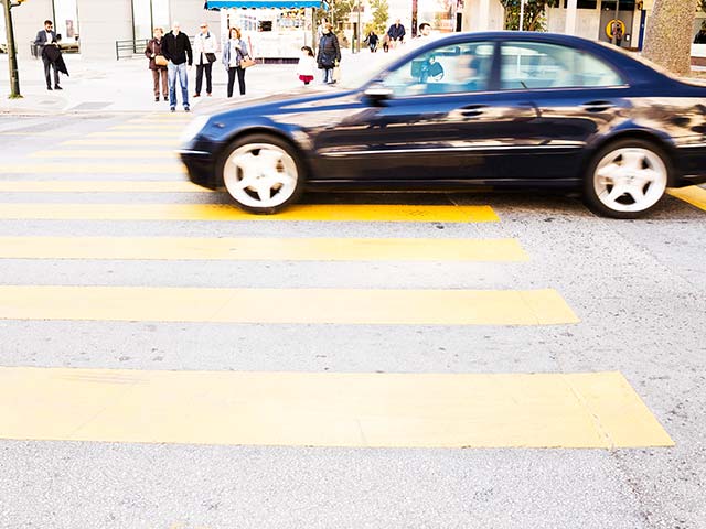 A black car speeds through a yellow pedestrian crossing on a city street, with people standing on the sidewalk.