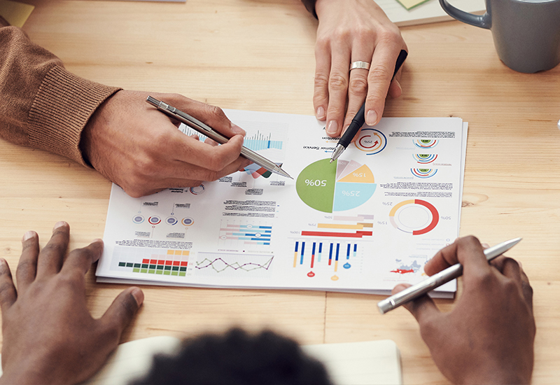 Hands holding pens over colorful charts and graphs on paper, discussing data.