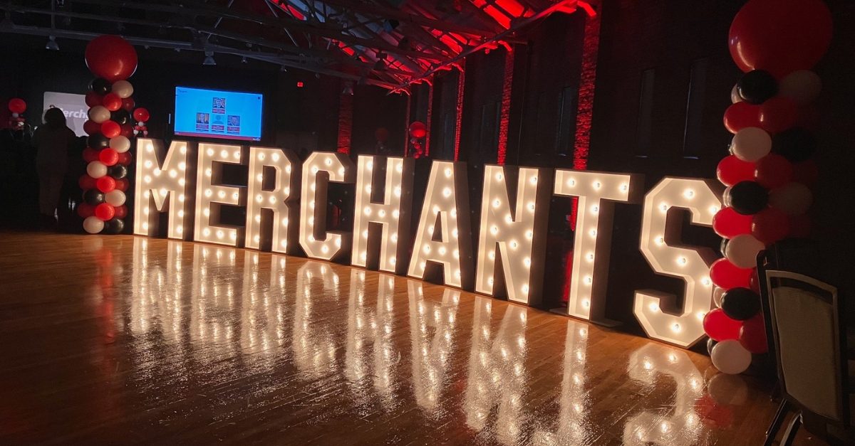 Large illuminated letters spell "MERCHANTS" amid red and black balloons in a dimly lit venue.