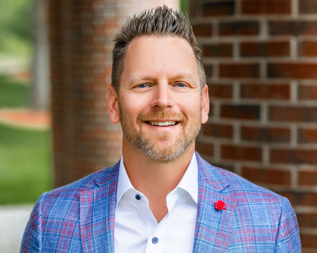 Smiling man in a blue plaid jacket with a red lapel pin, standing in front of a brick wall.