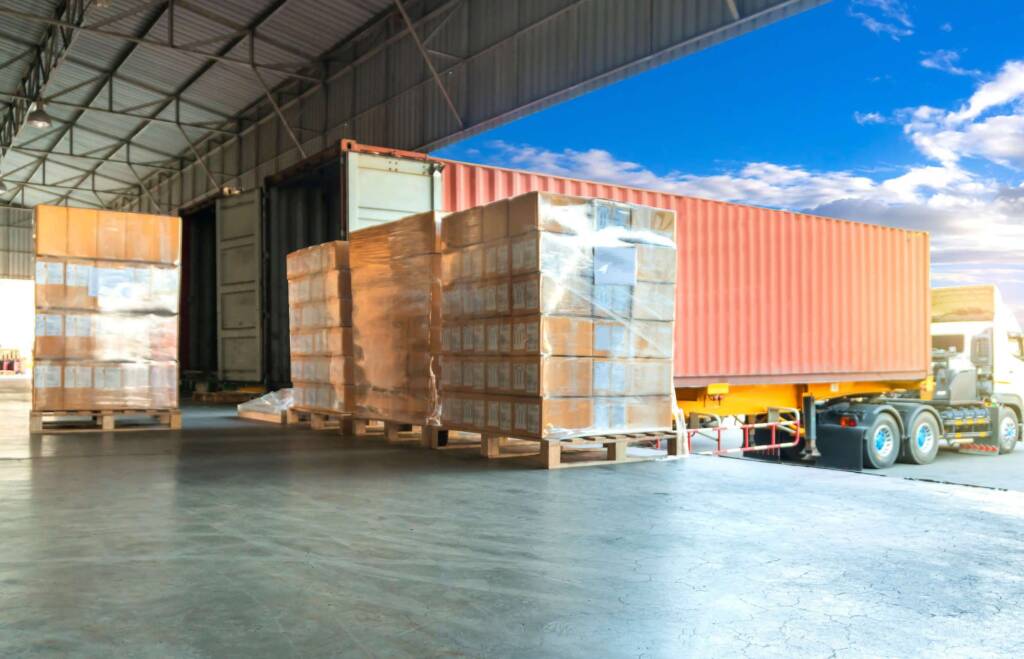Pallets of boxes wrapped in plastic are being loaded into an orange shipping container at a warehouse.