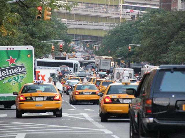 Busy city street with yellow taxis and various vehicles in traffic, surrounded by greenery and buildings.