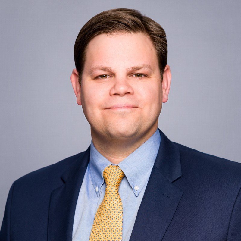 Smiling man in a blue suit and yellow tie, posing against a gray background.