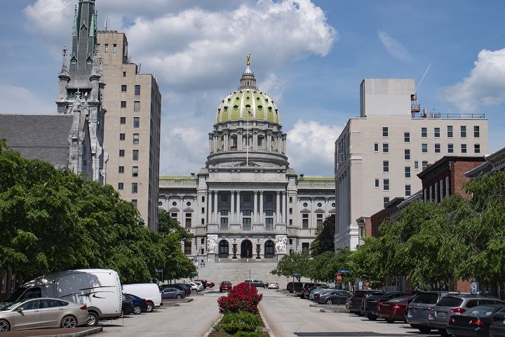 Street view of a domed capitol building flanked by trees and surrounded by tall buildings under a partly cloudy sky.