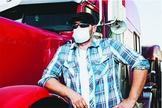 Person in a plaid shirt and face mask leans against a red truck, wearing a cap and sunglasses.
