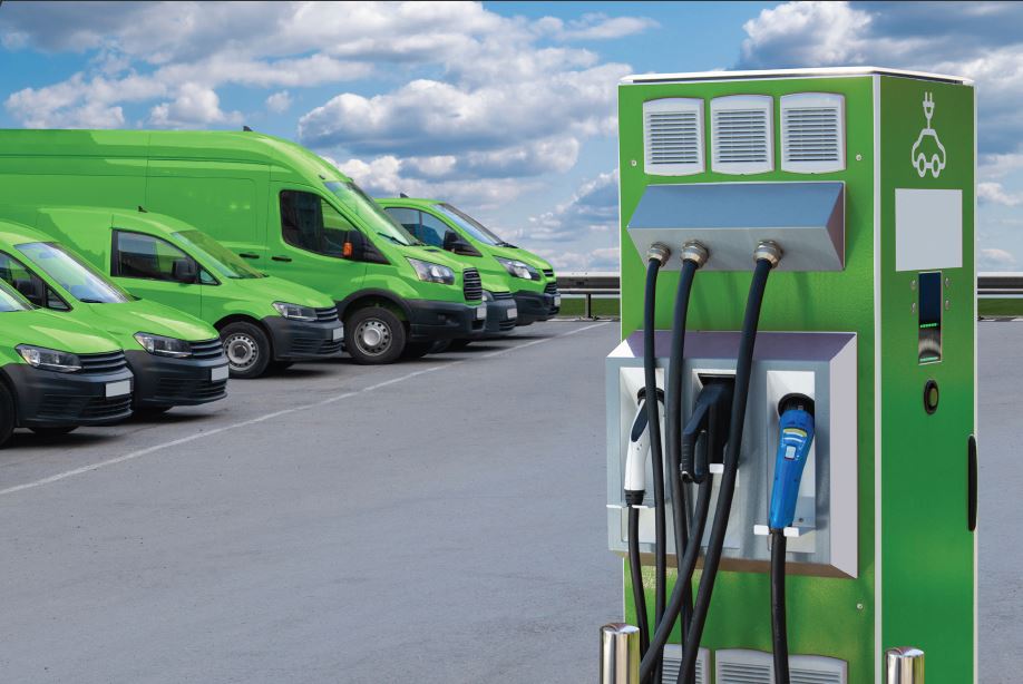 Green electric vans are parked near a charging station under a cloudy sky.