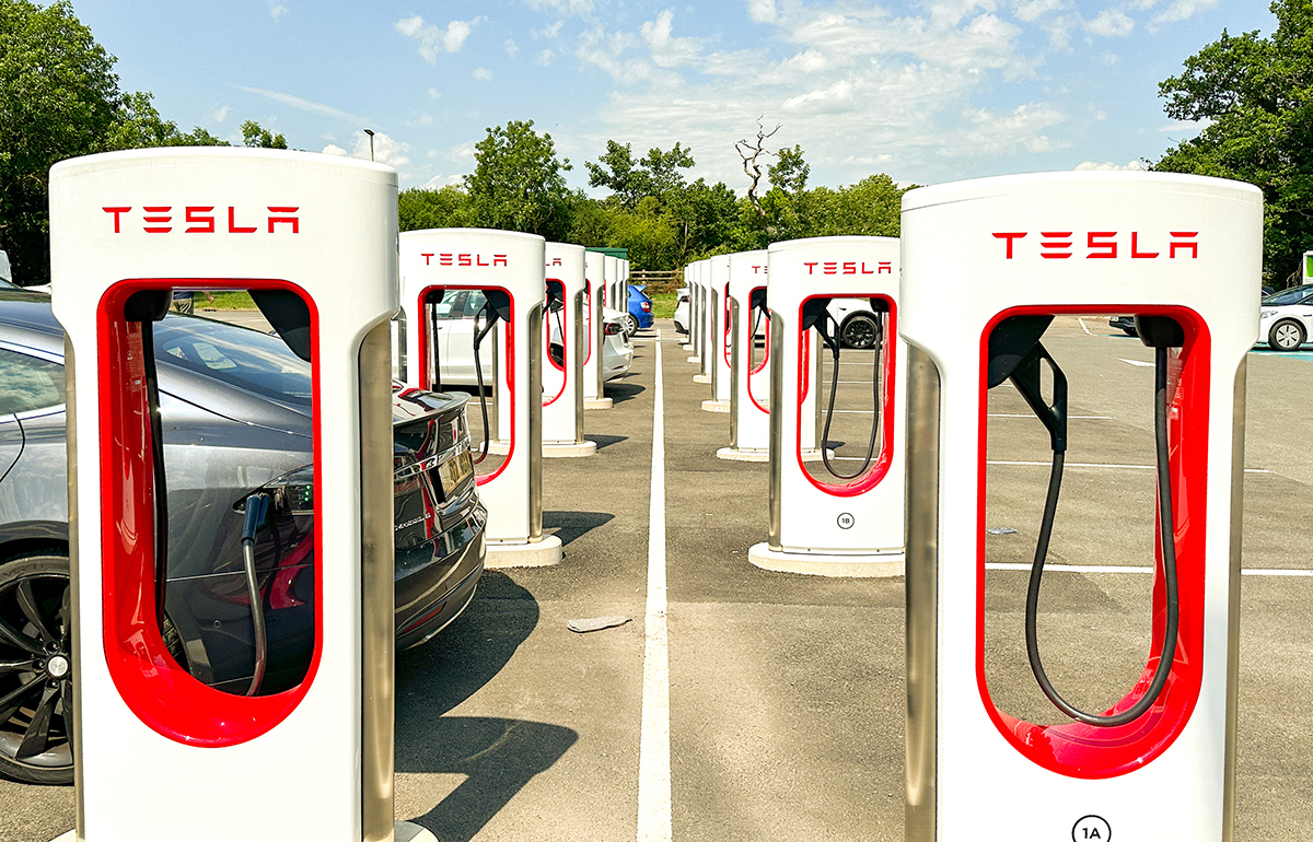 Tesla charging station with multiple chargers and parked cars in an outdoor parking lot beneath a clear sky.
