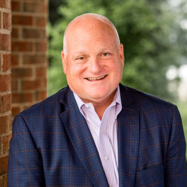 Smiling man in a checked suit jacket and striped shirt, standing outside near a brick wall with greenery in the background.
