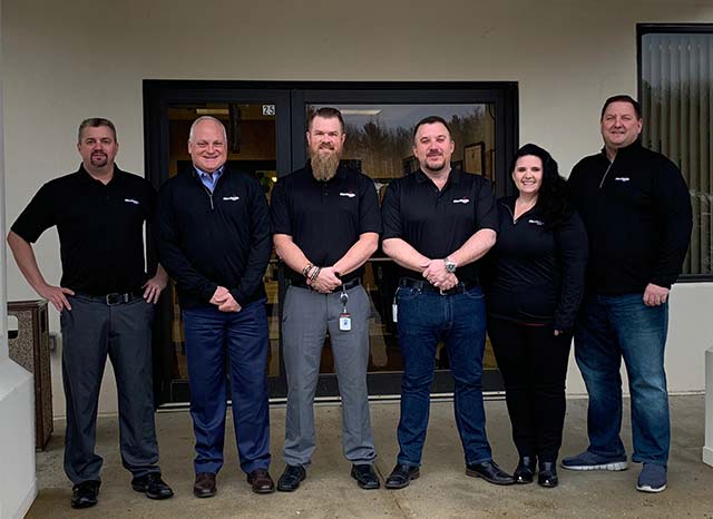 Six people in black shirts stand smiling outside a building entrance.