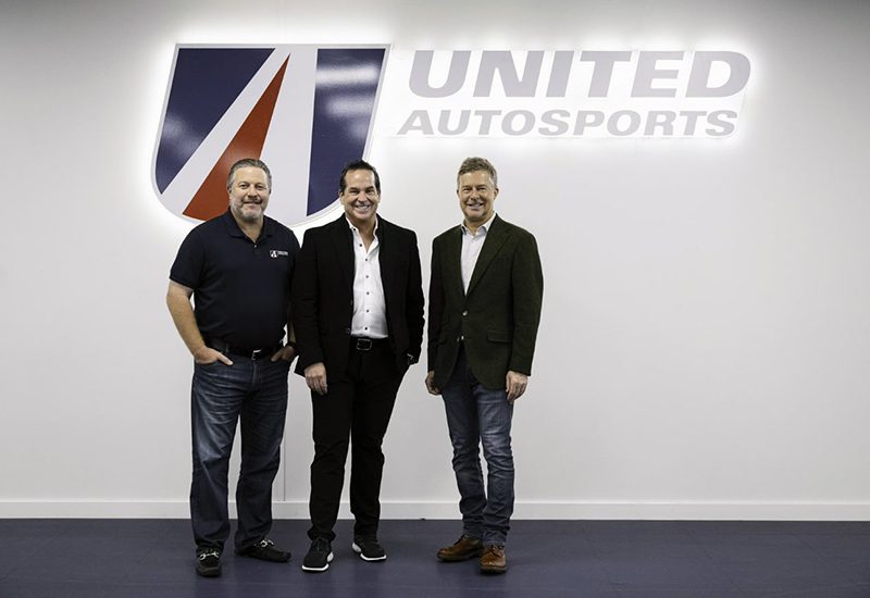 Three men standing in front of a United Autosports logo on a wall.