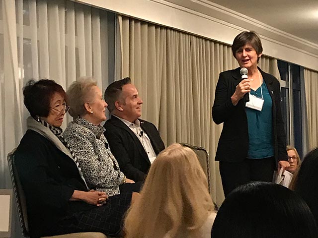 A woman speaks into a microphone while three people sit attentively on a panel in a conference room.