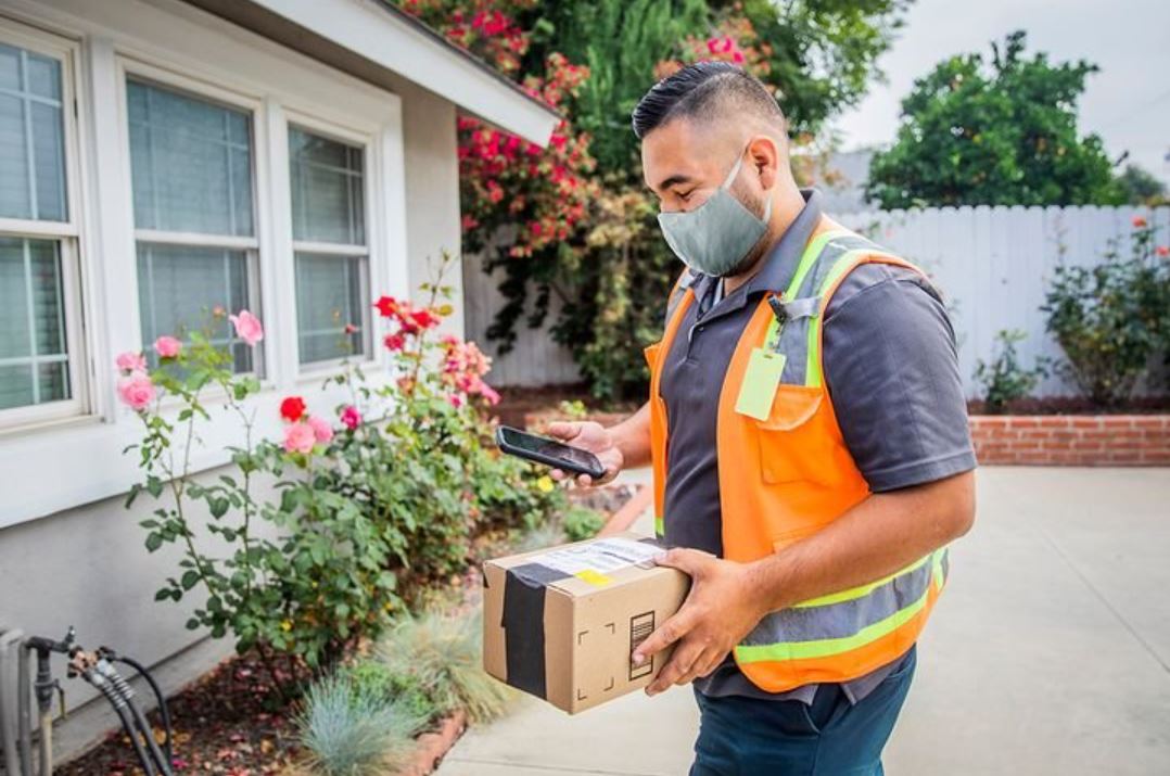 Delivery worker in orange vest scans a package in front of a house with flowers, wearing a gray mask.