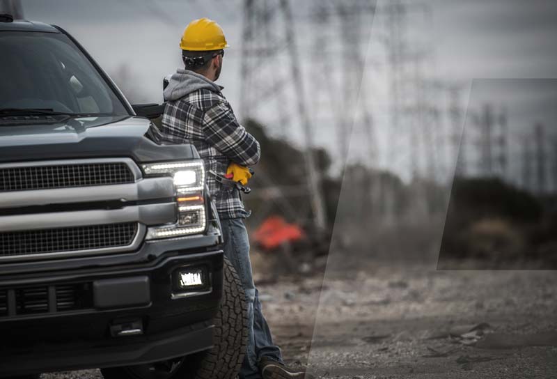 Person in a plaid jacket and yellow hard hat leans against a truck, looking at power lines in a rural area.