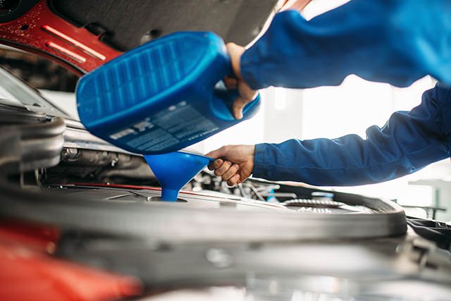 Mechanic pouring motor oil into a car engine using a blue container and funnel.