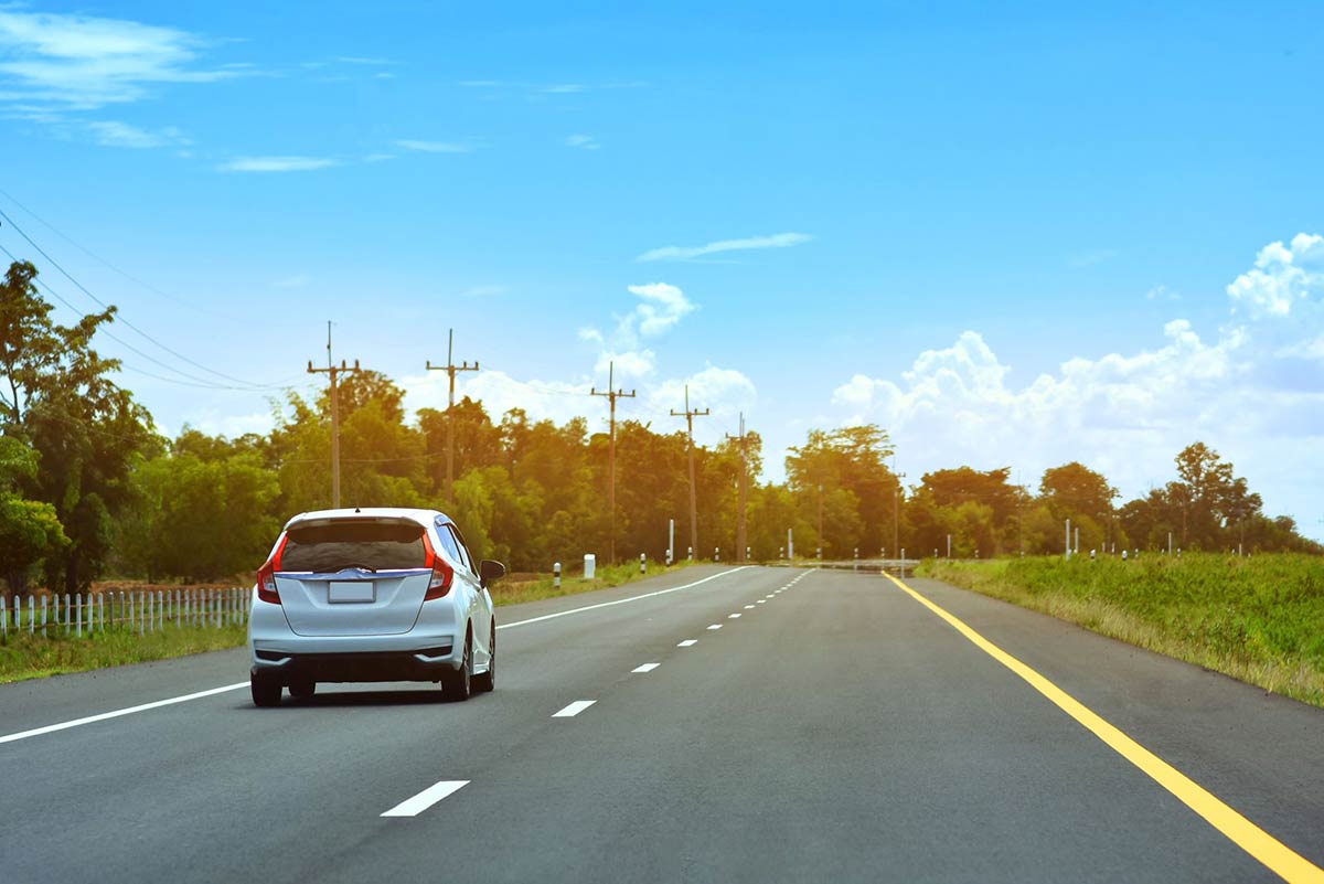 A white car driving on a clear rural road under a bright blue sky with scattered clouds and surrounding greenery.