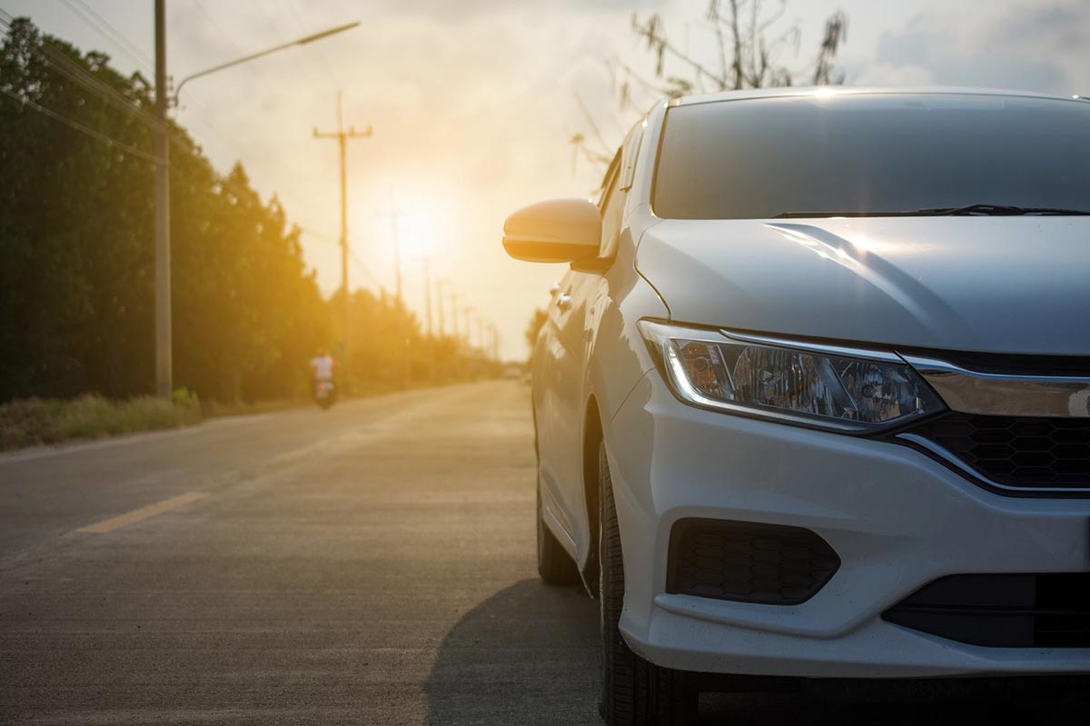 White car parked on a quiet road at sunset, with a motorcyclist in the distance.