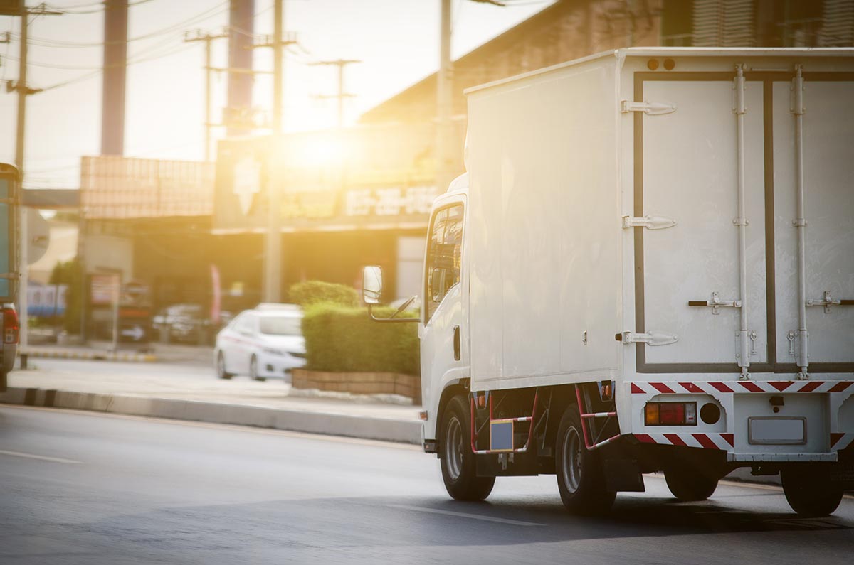 A delivery truck drives down a city street at sunset, with an industrial backdrop.