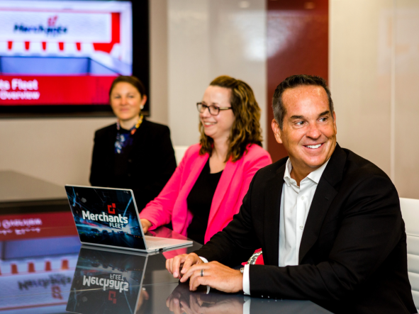 Three people in a meeting room, one with a laptop displaying "Merchants Fleet" on the screen.