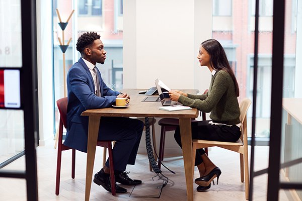 Two people sitting at a table in an office, engaged in a discussion, with laptops and documents on the table.
