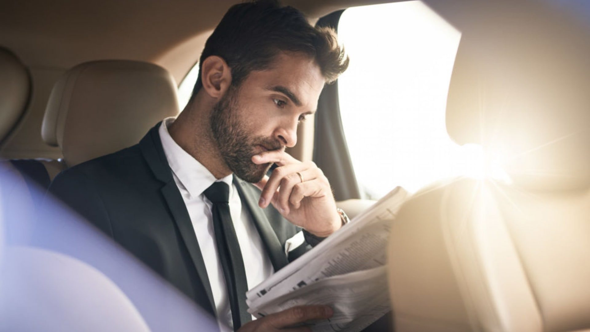 Man in a suit reading a newspaper in the backseat of a car, with sunlight filtering through the window.