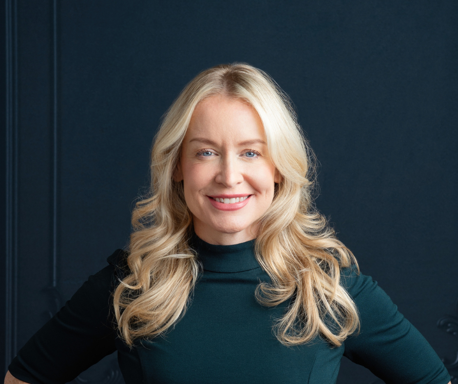 Person with long blonde hair and a dark top smiling against a dark background.