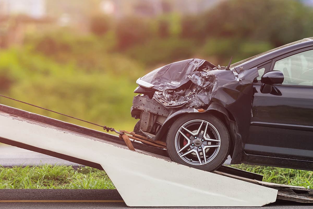 A damaged black car being loaded onto a tow truck ramp with a grassy background.