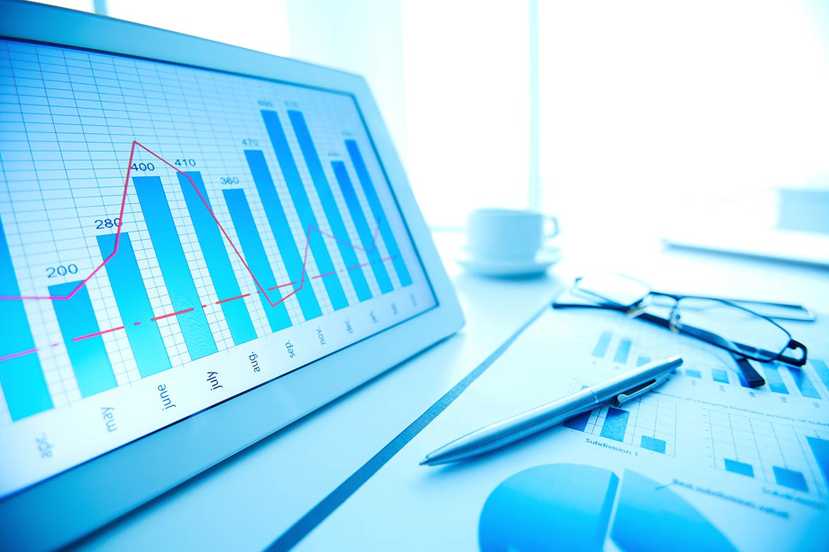 Tablet showing bar and line graphs on a desk with papers, a pen, glasses, and a coffee cup.