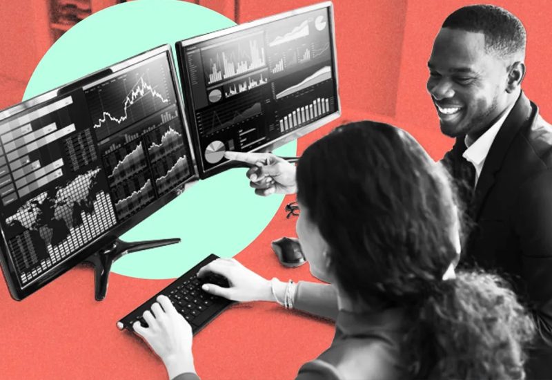 Two people analyze financial graphs on dual computer monitors in an office setting.