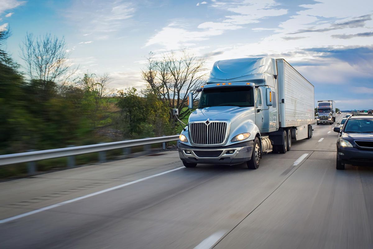 Silver semi-truck driving on a highway with other vehicles, trees and a cloudy sky in the background.