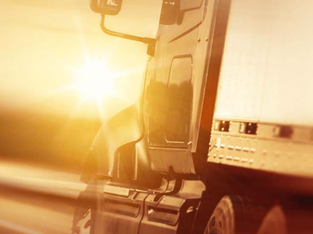 Semi-truck on highway at sunset, with sun glaring on the side of the cab and trailer.