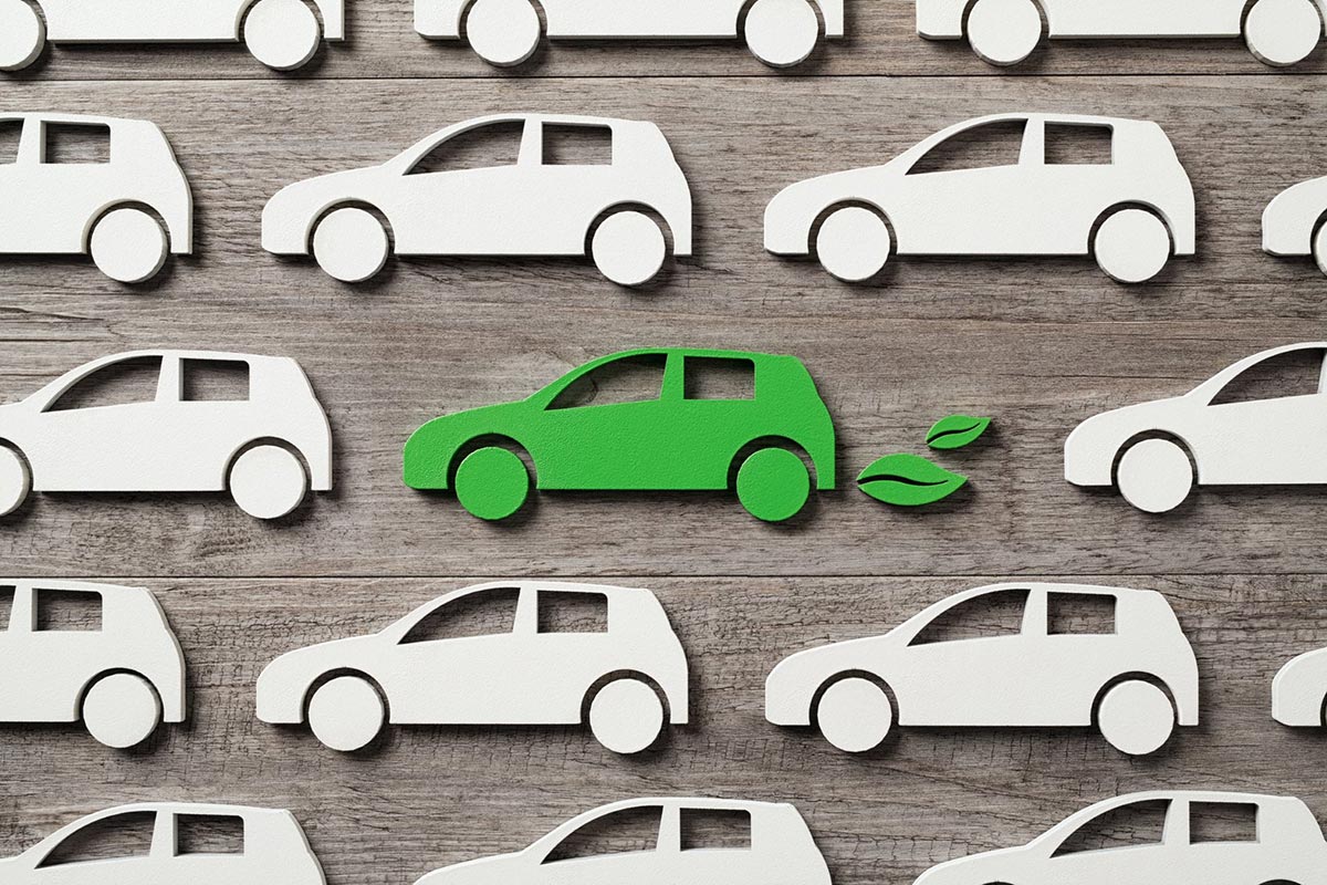 Pattern of white car cutouts on wood, with one green car and leaf cutouts, symbolizing eco-friendly vehicles.