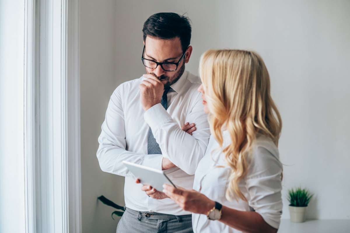 Two people in a white office, one holding a tablet, both looking thoughtful near a window.