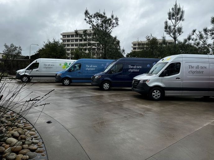 Four Mercedes-Benz eSprinter vans parked on wet pavement, surrounded by trees and buildings under a cloudy sky.