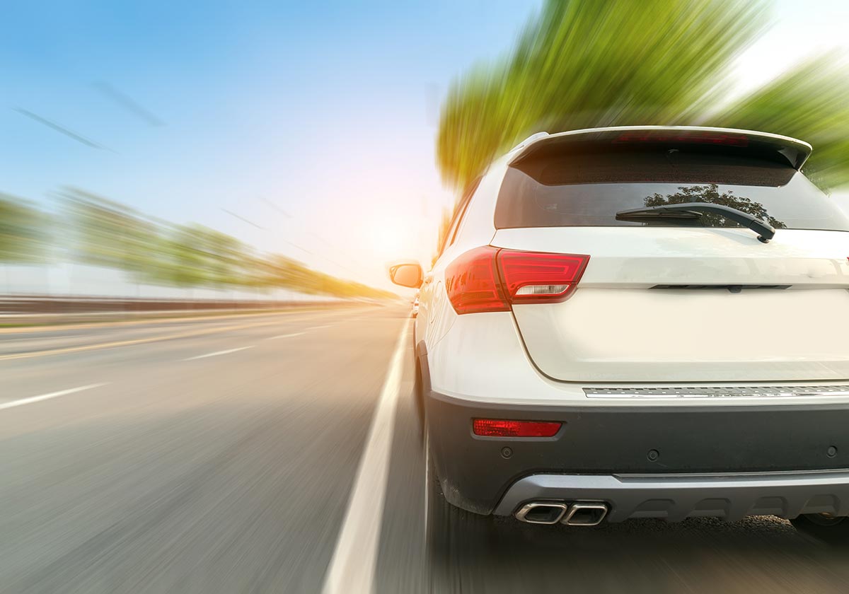 White SUV driving fast on a highway with blurred trees and a bright sky in the background.