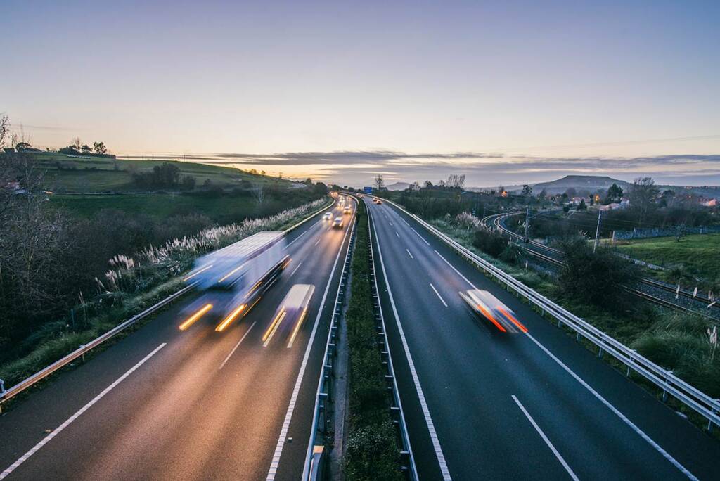 Blurred cars drive on a dual carriageway at dusk, with soft hills and distant lights under a twilight sky.