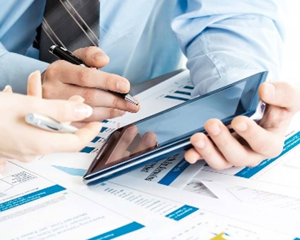 Hands of two people discussing and pointing at a tablet over scattered financial documents.