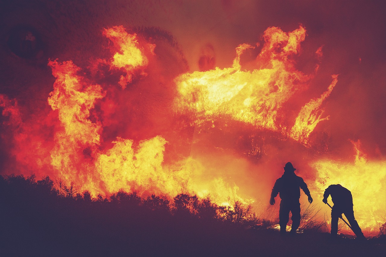 Silhouetted firefighters battle a large, intense brush fire at night, with bright orange flames in the background.